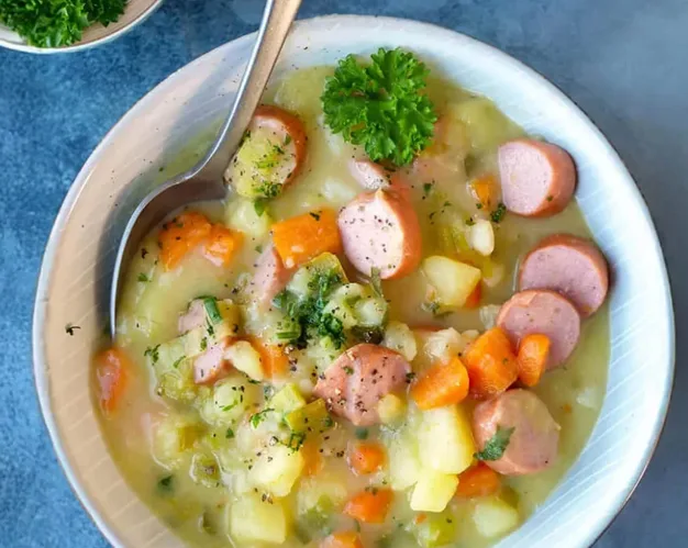 Kartoffelsuppe mit Würstchenscheiben, Kartoffel- und Möhrenstücken und grünen Kräutern in einer Schale.
